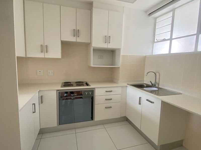 A clean, modern kitchen featuring white cabinetry, built-in oven and hob, stainless steel sink, and neutral tiled flooring.