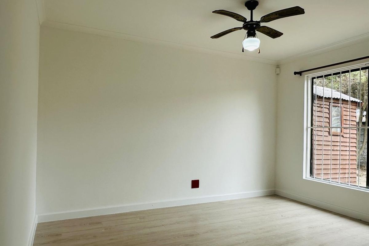 A bright, empty bedroom featuring light-toned laminate flooring, a ceiling fan, and a window fitted with security bars.