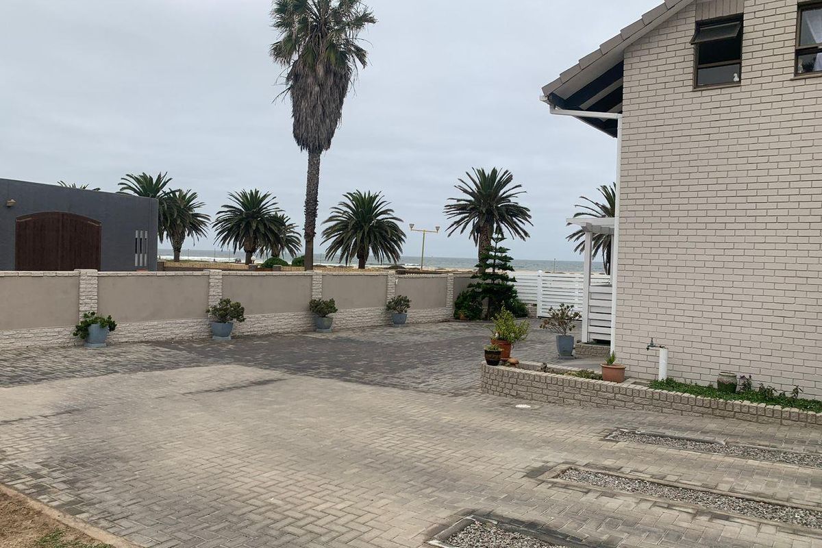 A paved driveway and courtyard area featuring brick boundary walls, established palm trees, and a view towards the ocean.