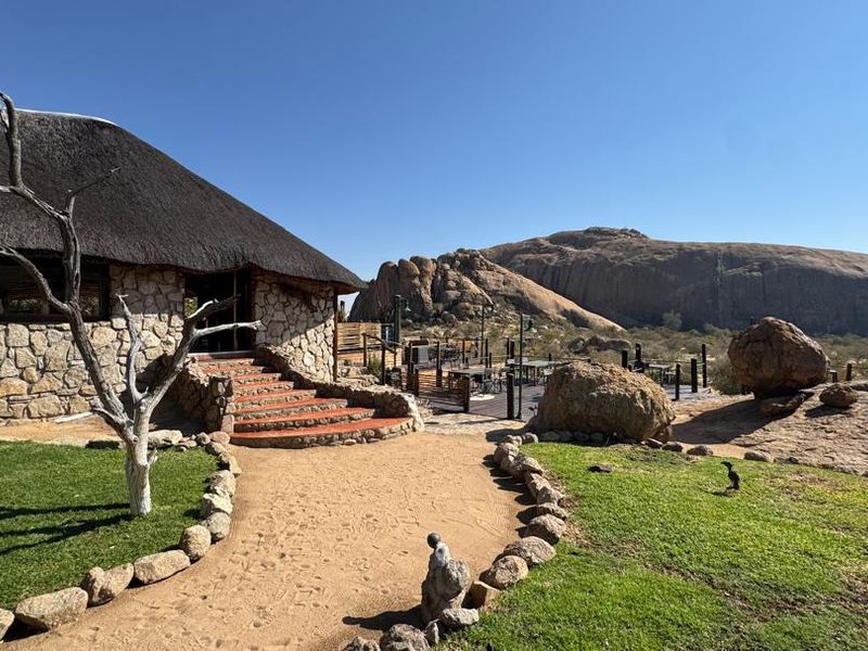 A scenic view of a lodge exterior featuring traditional thatched roofing, stone masonry, and a landscaped garden set against a dramatic rocky mountain backdrop.