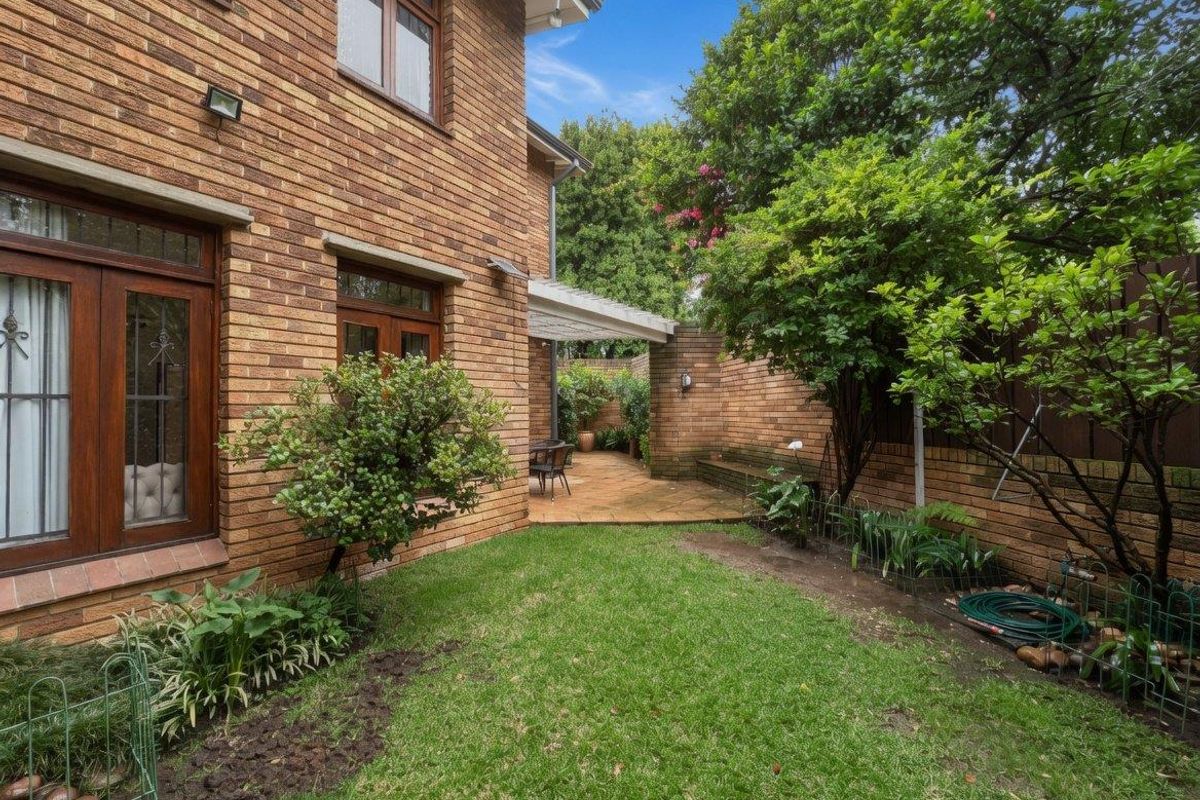 A private rear garden featuring a brick townhouse facade, a paved patio area with a pergola, and established greenery.