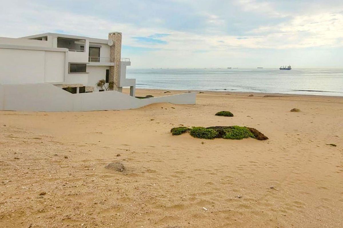 A modern residential property situated directly on the beachfront, featuring a stone chimney and balcony with expansive ocean views.