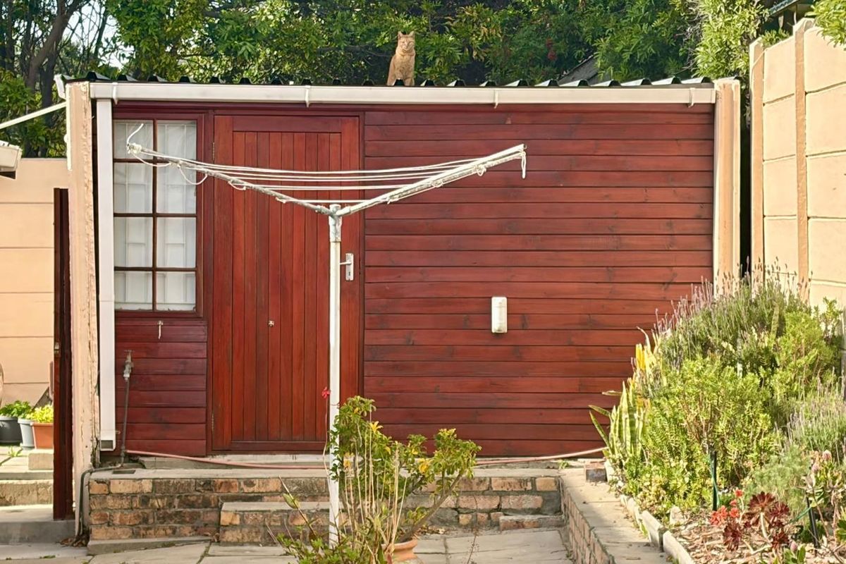 A rear exterior view of a timber-clad structure featuring a wooden door, window, and paved patio area with a brick retaining wall.