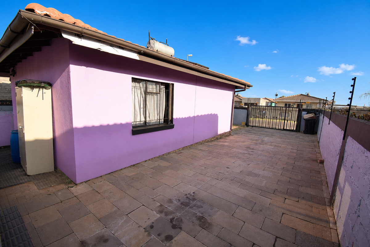 Rear exterior view featuring a paved courtyard, security gate, perimeter wall with electric fencing, and a solar geyser installed on the tiled roof.