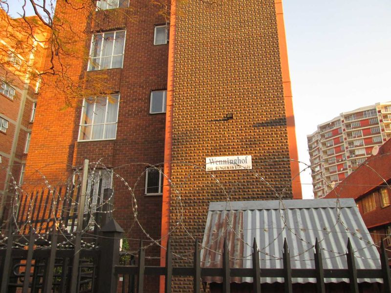 Exterior view of the Wenninghof apartment building, featuring a brick facade, secure perimeter fencing with razor wire, and a metal entrance structure.