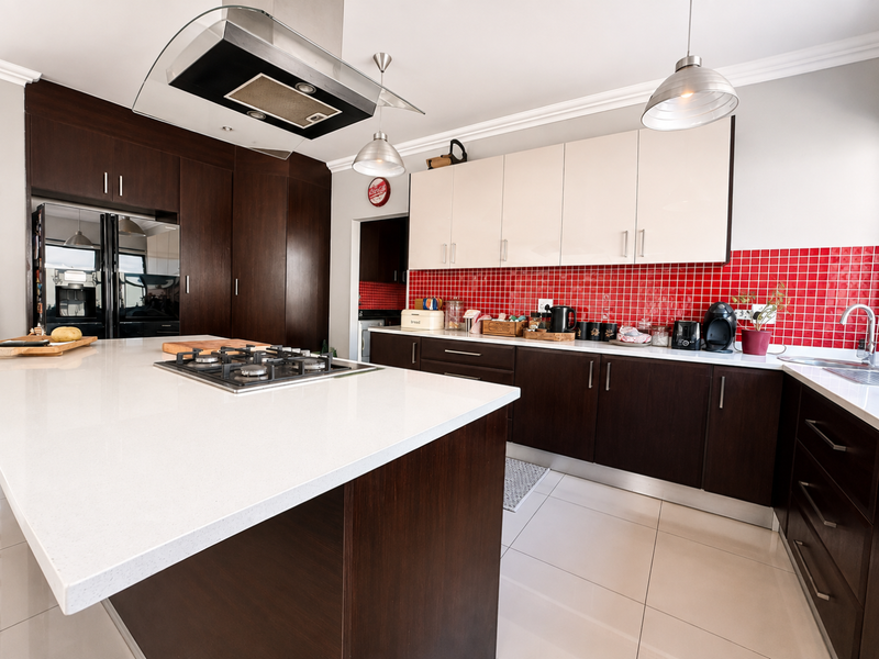 Modern kitchen featuring a large island with gas hob, contrasting dark and light cabinetry, stone countertops, and a vibrant red tiled backsplash.