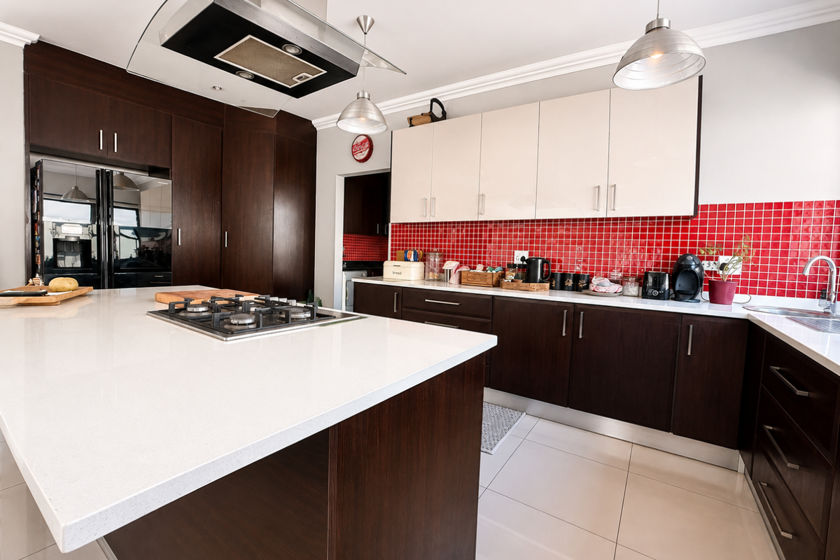 Modern kitchen featuring a large island with gas hob, contrasting dark and light cabinetry, stone countertops, and a vibrant red tiled backsplash.