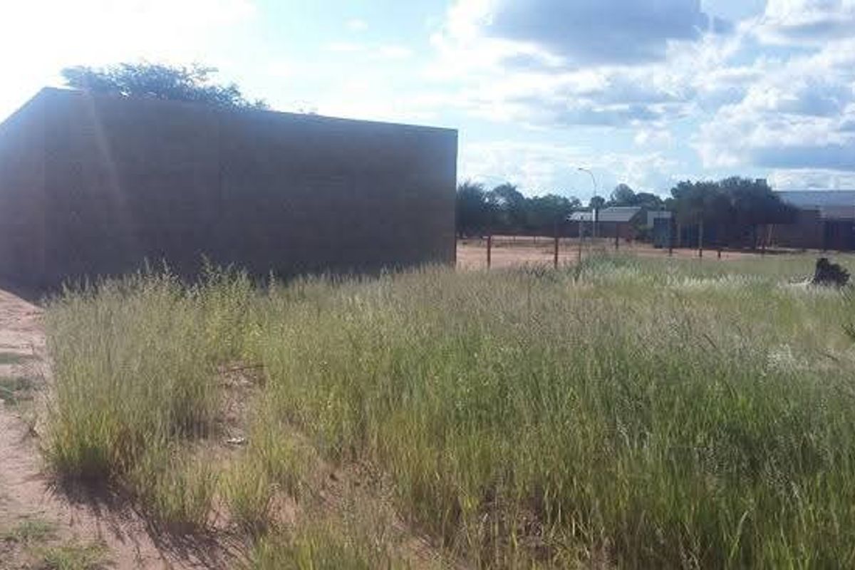 A view of a vacant commercial plot featuring a boundary wall and overgrown grass, situated in a developing area.