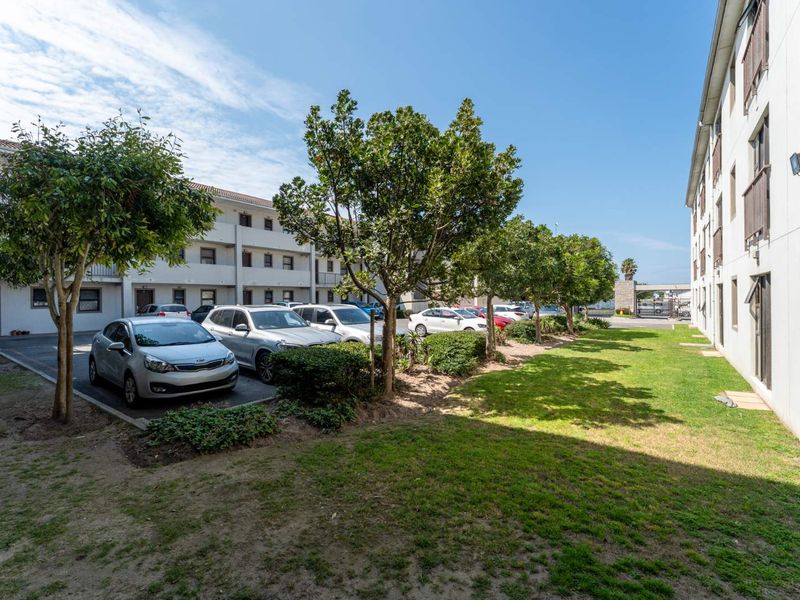 Exterior view of a multi-story residential complex featuring a paved parking area, landscaped garden beds, and established trees between building blocks.