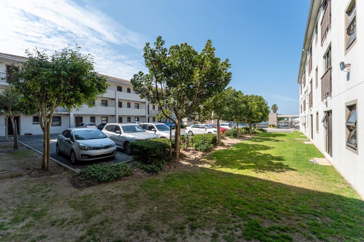 Exterior view of a multi-story residential complex featuring a paved parking area, landscaped garden beds, and established trees between building blocks.