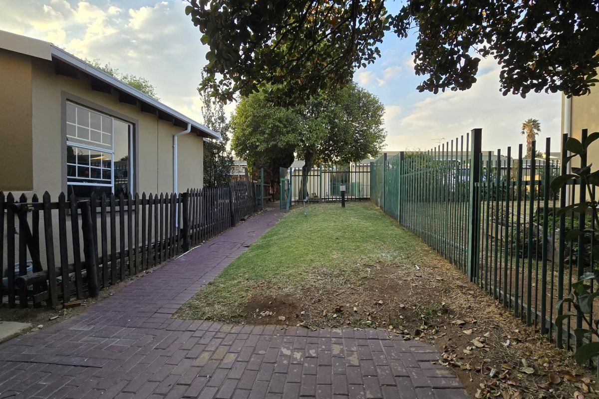 A rear exterior view of the townhouse featuring a paved walkway, a small lawn area, and secure perimeter fencing.