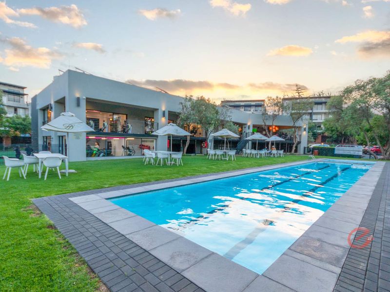 A well-maintained communal pool area featuring a paved surround, manicured lawn, and views of the adjacent clubhouse and gym facility.