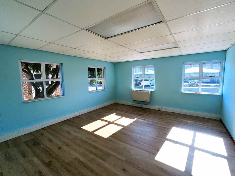 A bright, empty office space featuring laminate flooring, a suspended ceiling, and an integrated air conditioning unit with multiple windows providing natural light.