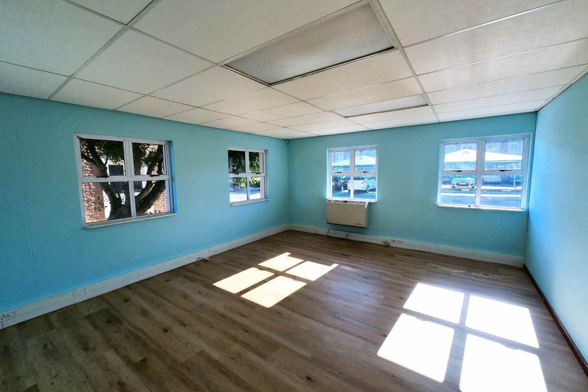 A bright, empty office space featuring laminate flooring, a suspended ceiling, and an integrated air conditioning unit with multiple windows providing natural light.