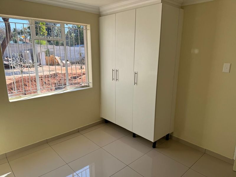 A clean, empty bedroom featuring neutral tiled flooring, a built-in wardrobe, and a window fitted with security bars.