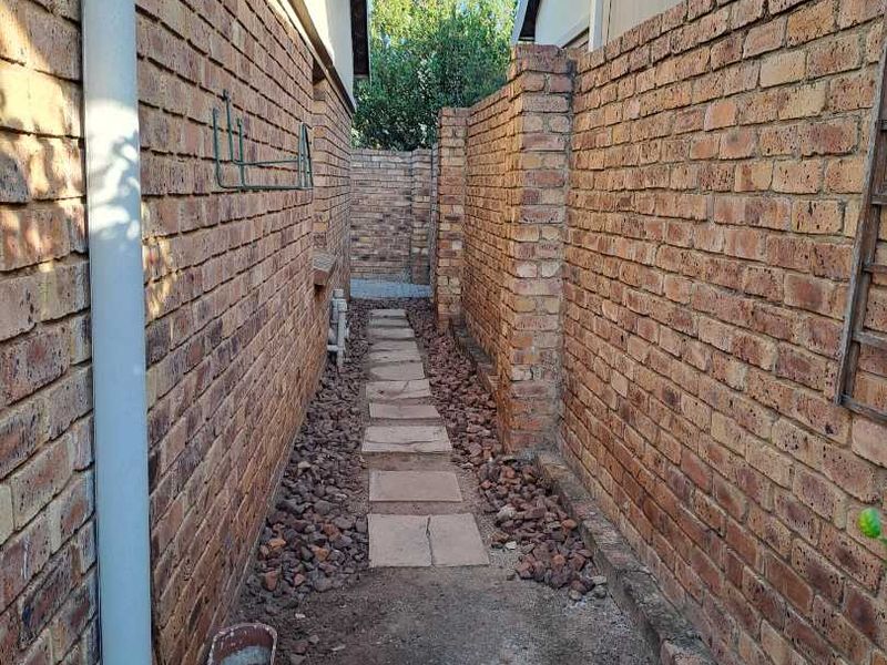 A narrow side passage between two brick buildings featuring a paved walkway, gravel ground cover, and brick boundary walls.