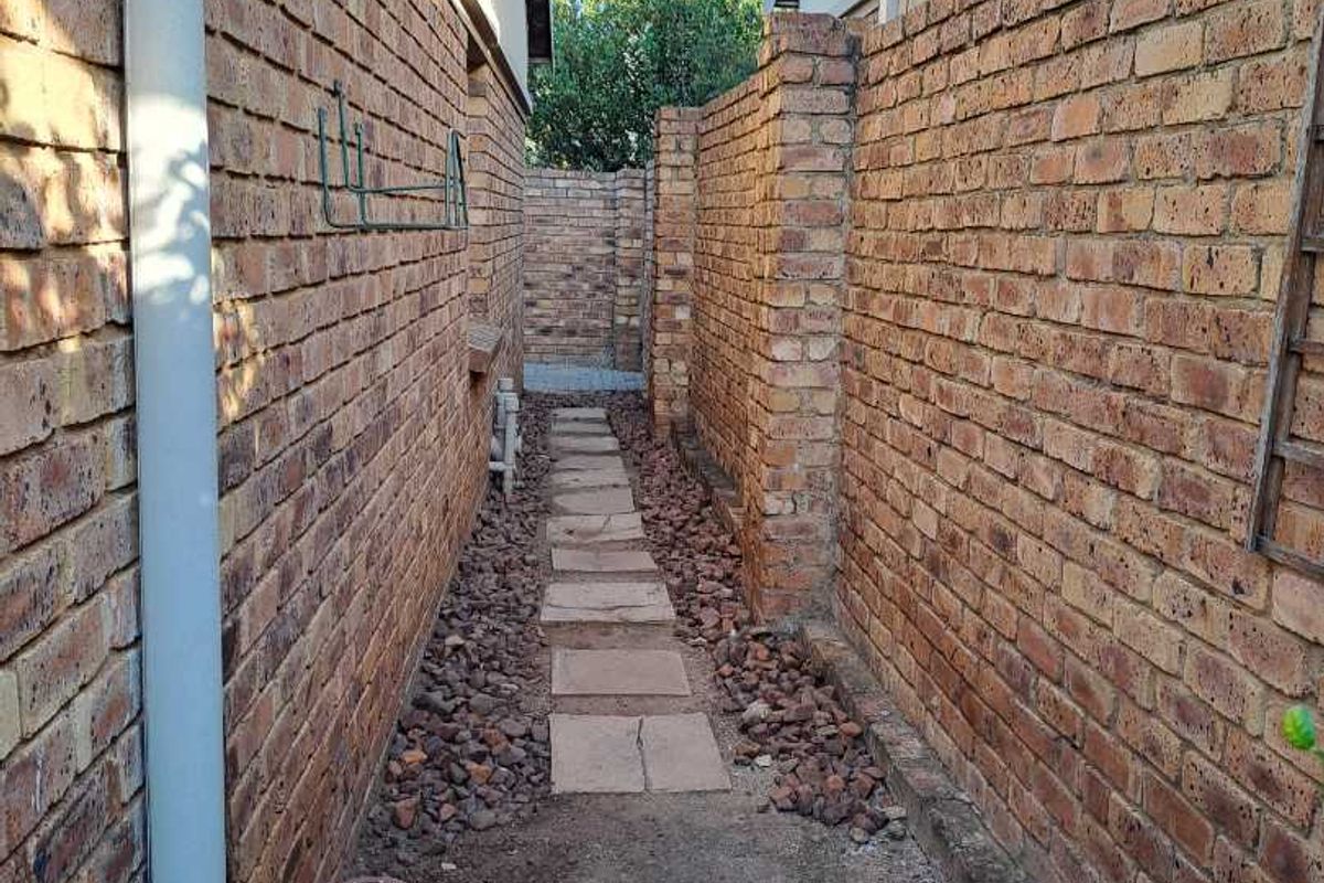 A narrow side passage between two brick buildings featuring a paved walkway, gravel ground cover, and brick boundary walls.