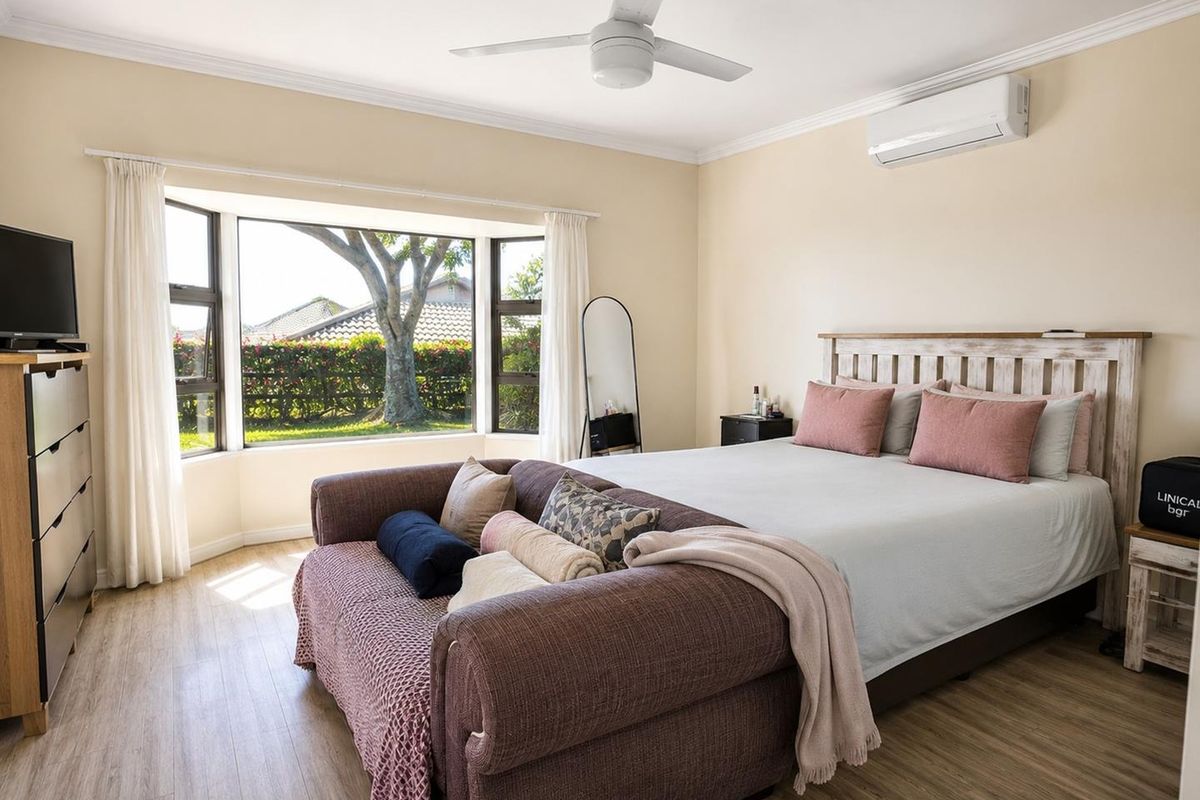 A well-maintained bedroom featuring laminate flooring, a ceiling fan, a split-system air conditioner, and a large bay window overlooking the garden.