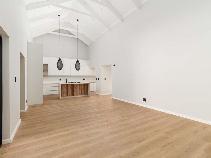 Spacious open-plan living area featuring a vaulted ceiling with exposed rafters, modern kitchen cabinetry, and light wood-look flooring.