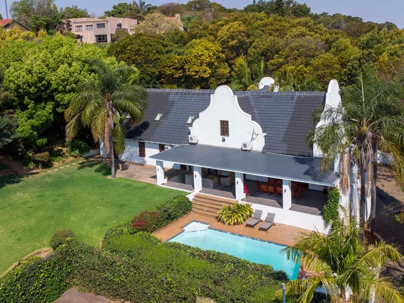 Aerial view of a Cape Dutch style home featuring a swimming pool, expansive covered patio, and lush landscaped garden surroundings.