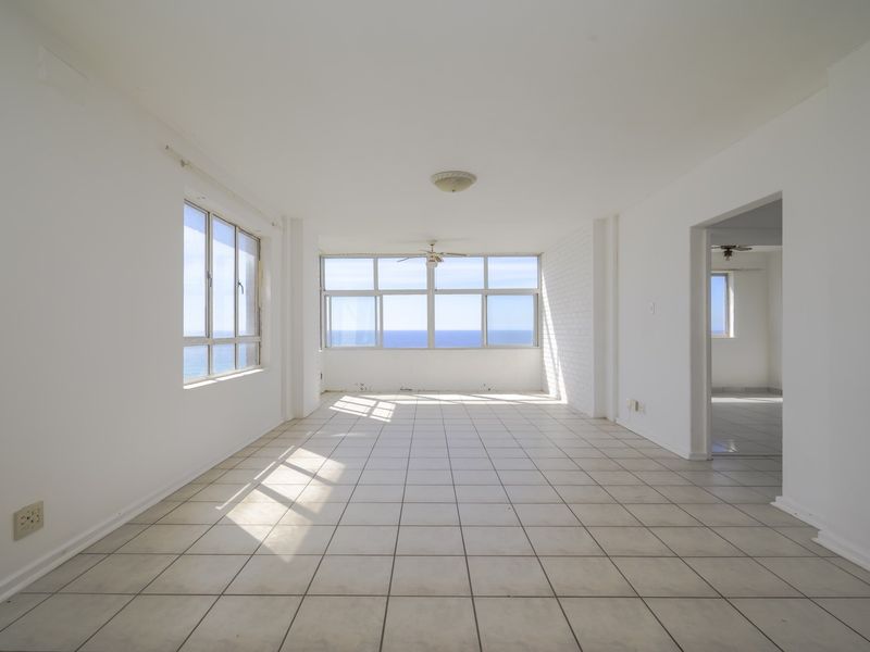 A bright, empty living area featuring tiled flooring, a ceiling fan, and expansive windows offering panoramic ocean views.