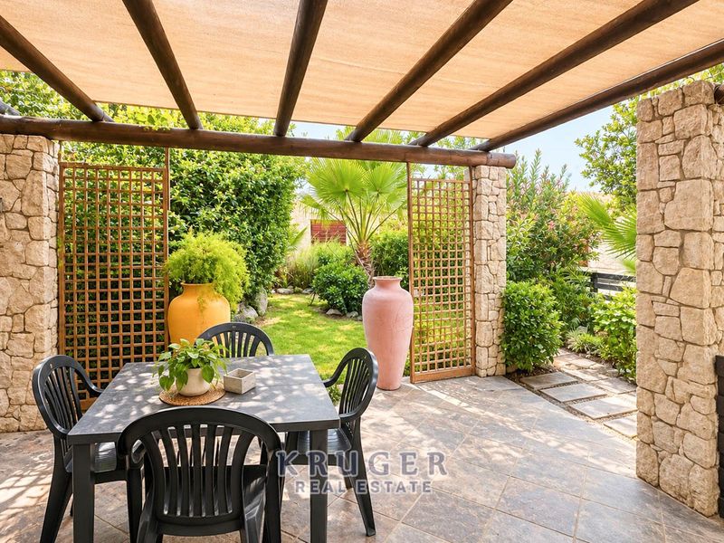 A covered outdoor patio featuring stone pillars, a timber pergola with shade cloth, and tiled flooring, overlooking a landscaped garden area.