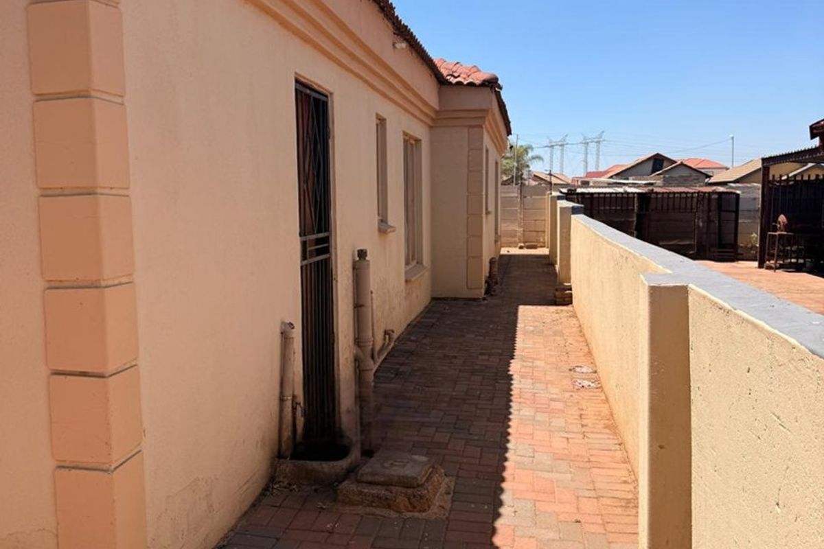 A side exterior view of the property featuring a paved walkway, boundary wall, and external plumbing fixtures along the building facade.
