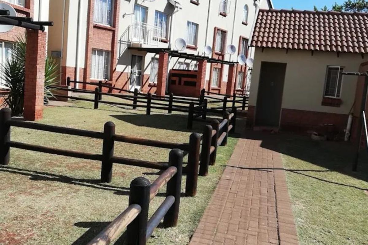 Exterior view of the apartment complex featuring a paved walkway, landscaped lawn, and timber fencing leading to the residential units.