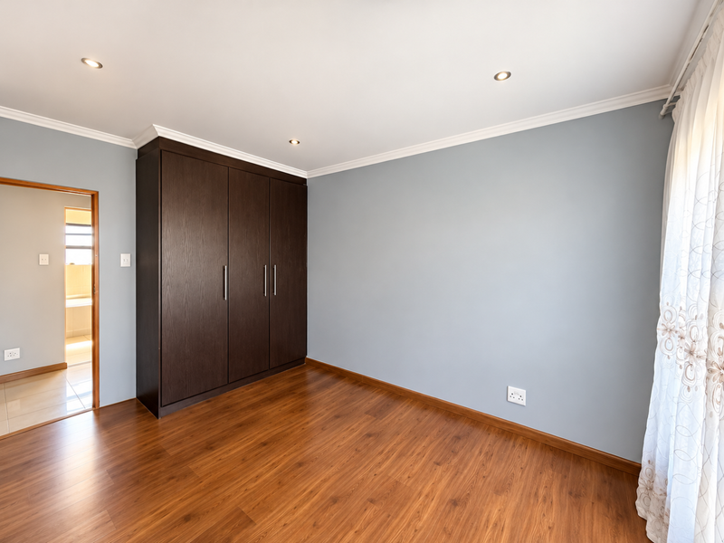 A bright, empty bedroom featuring dark wood-toned laminate flooring, a large built-in wardrobe, and recessed ceiling lighting.