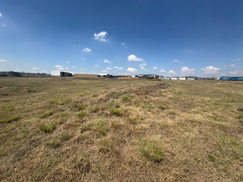 A wide-angle view of an undeveloped commercial land plot featuring open grassland with surrounding industrial buildings in the distance.