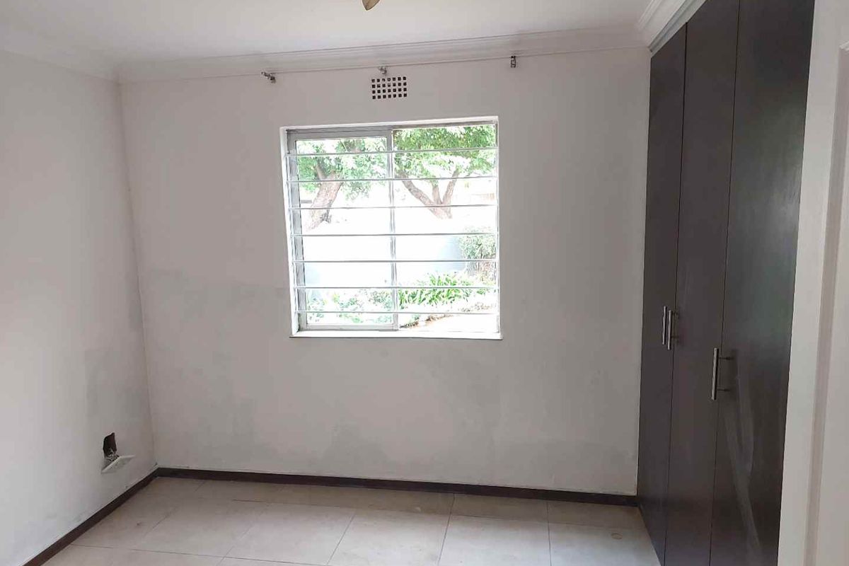 An empty bedroom featuring tiled flooring, built-in cupboards, and a window with security bars, offering a blank canvas for personalization.