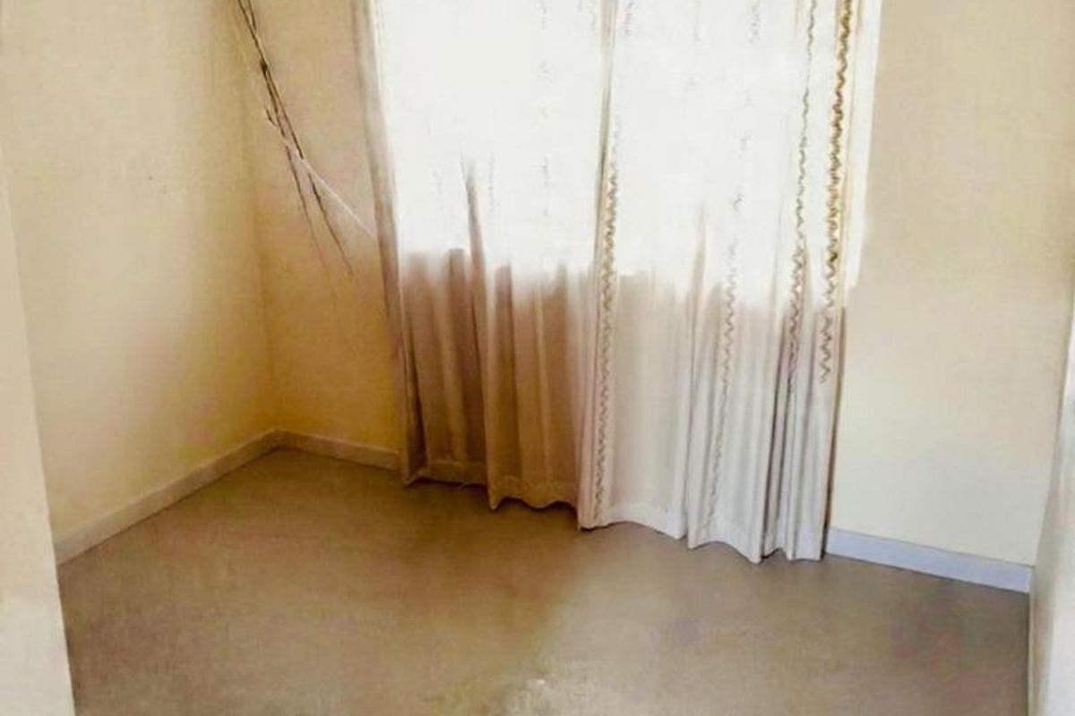 An empty bedroom featuring plain walls, a window with curtains, and visible floor staining. The room requires cosmetic updates and electrical repairs.