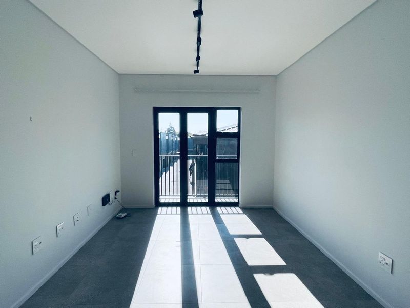 A bright, empty living room featuring modern grey floor tiles, track lighting, and large black-framed glass doors leading to an outdoor area.