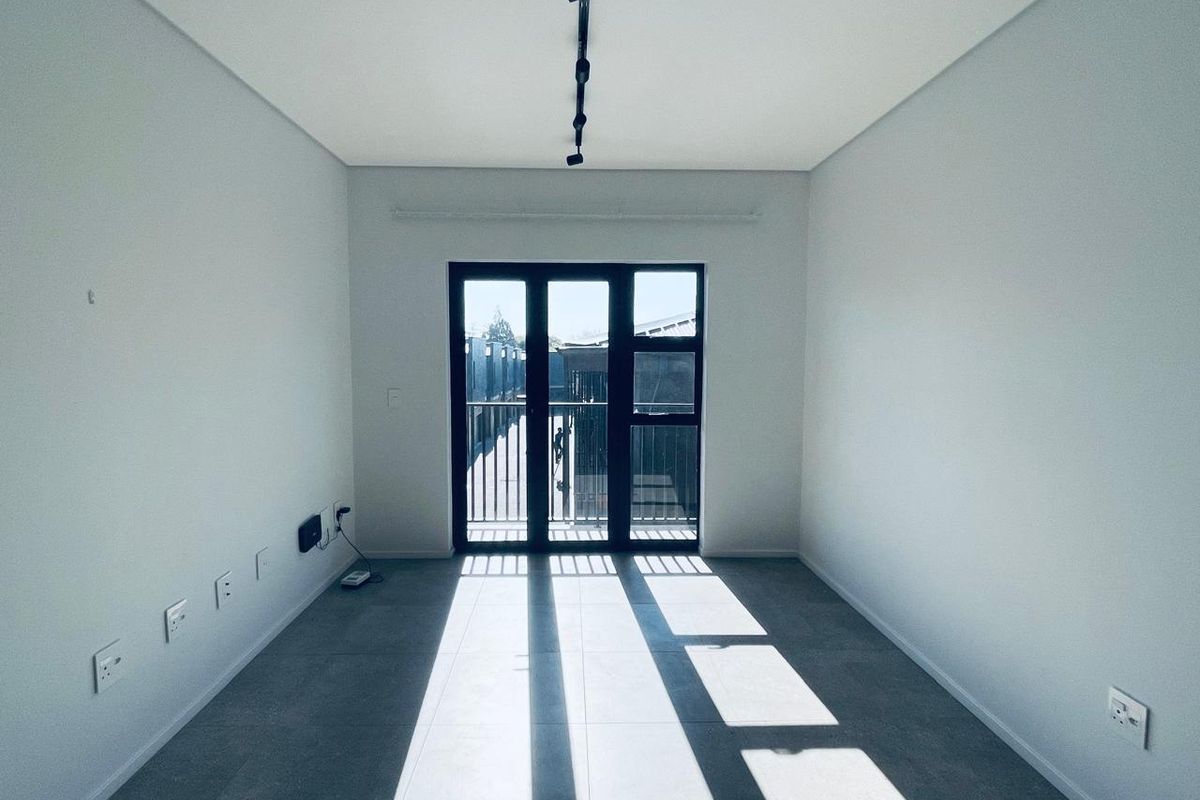 A bright, empty living room featuring modern grey floor tiles, track lighting, and large black-framed glass doors leading to an outdoor area.