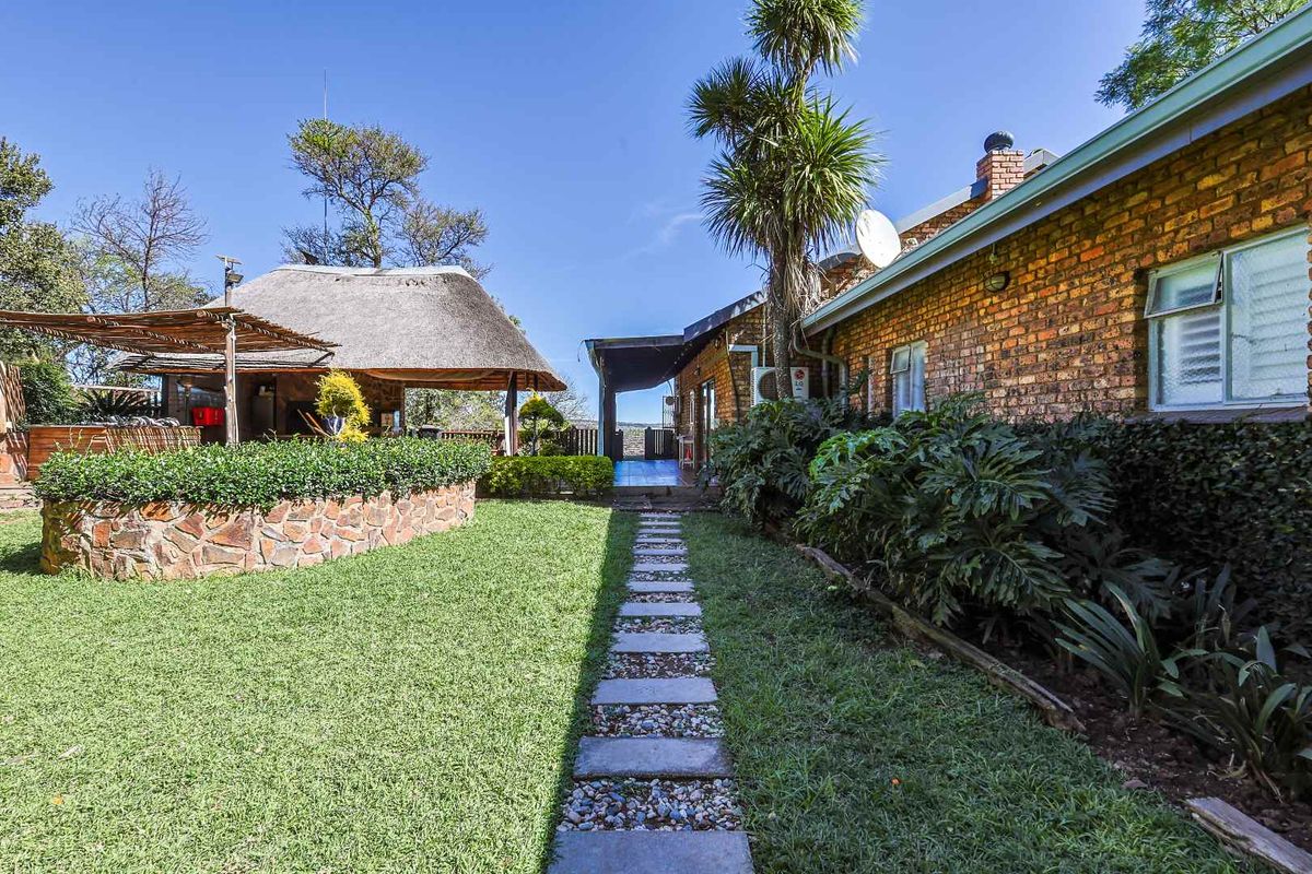 A well-maintained rear garden featuring a stone-clad raised planter, a thatched lapa entertainment area, and a paved walkway leading to the main residence.