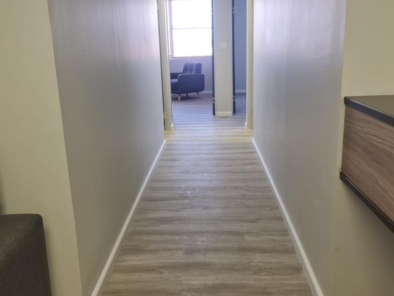 A clean, well-lit office hallway featuring modern wood-look vinyl flooring, neutral walls, and a drop ceiling with integrated lighting.