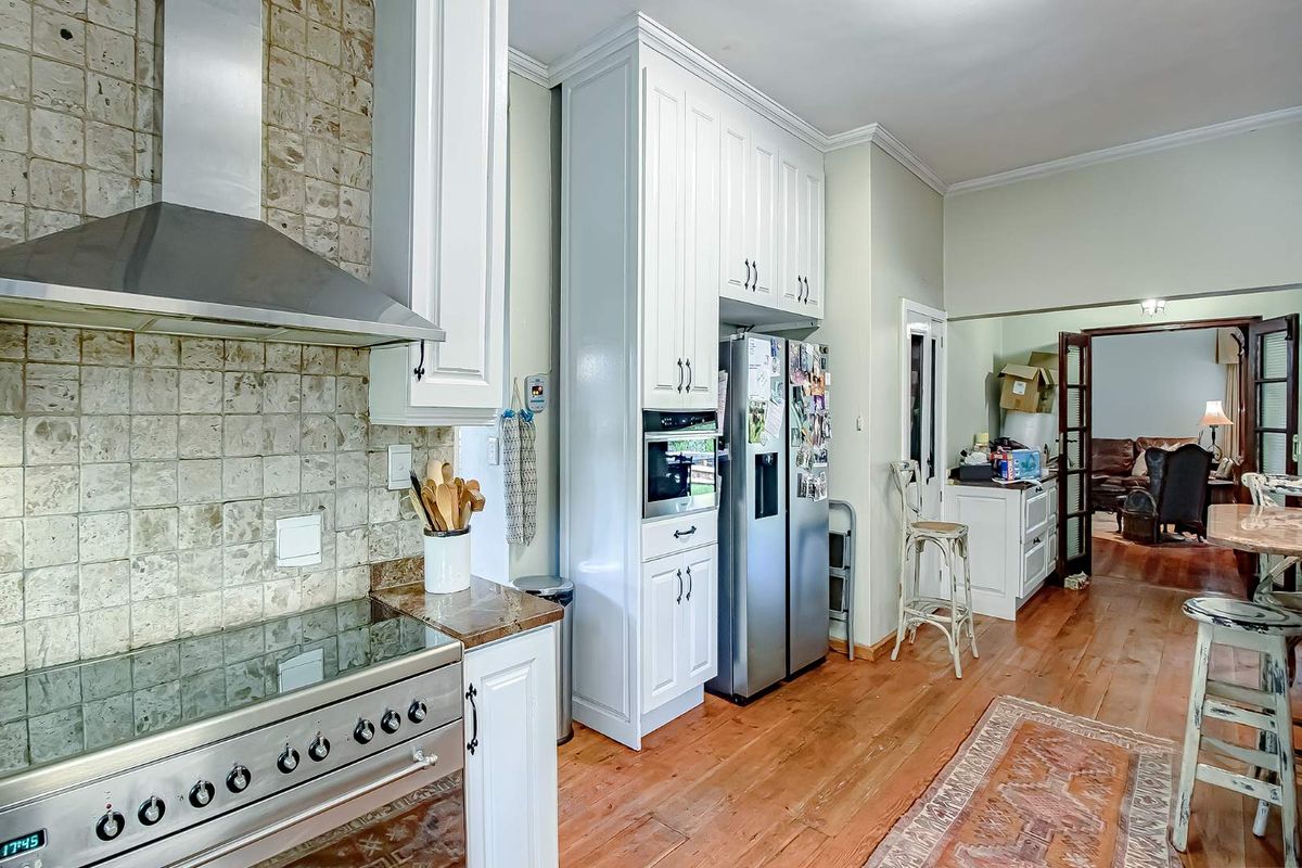 A well-appointed kitchen featuring white cabinetry, a stainless steel oven with extractor fan, stone-tiled backsplash, and warm wooden flooring, leading into an adjacent living area.