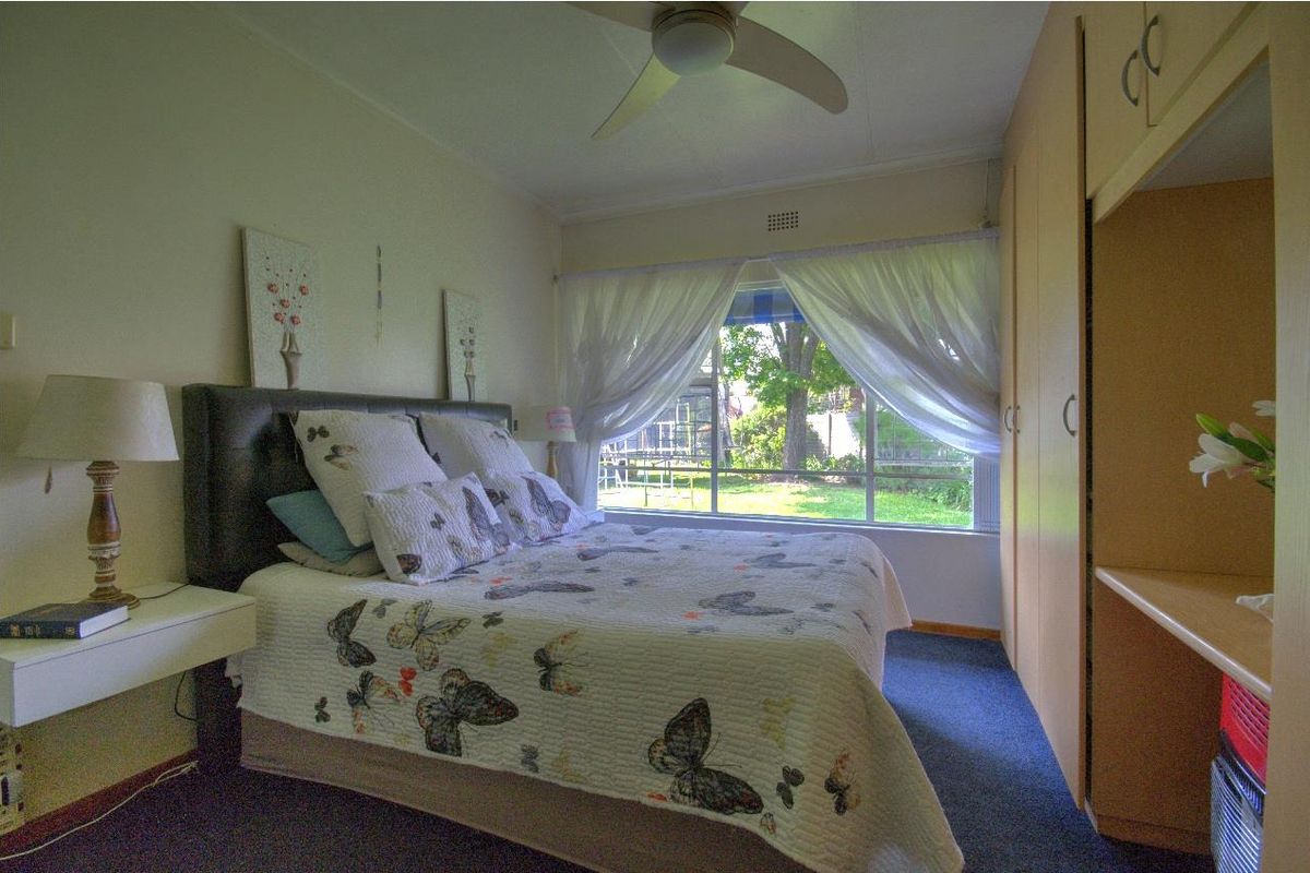 A functional bedroom featuring built-in cupboards, a ceiling fan, and a large window providing natural light and garden views.