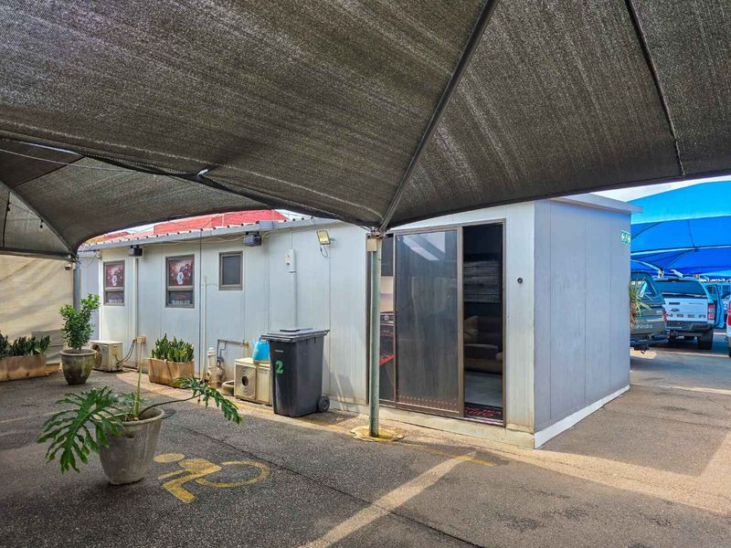 Exterior view of a commercial office unit featuring shade netting, asphalt parking area, and a sliding glass entrance door.