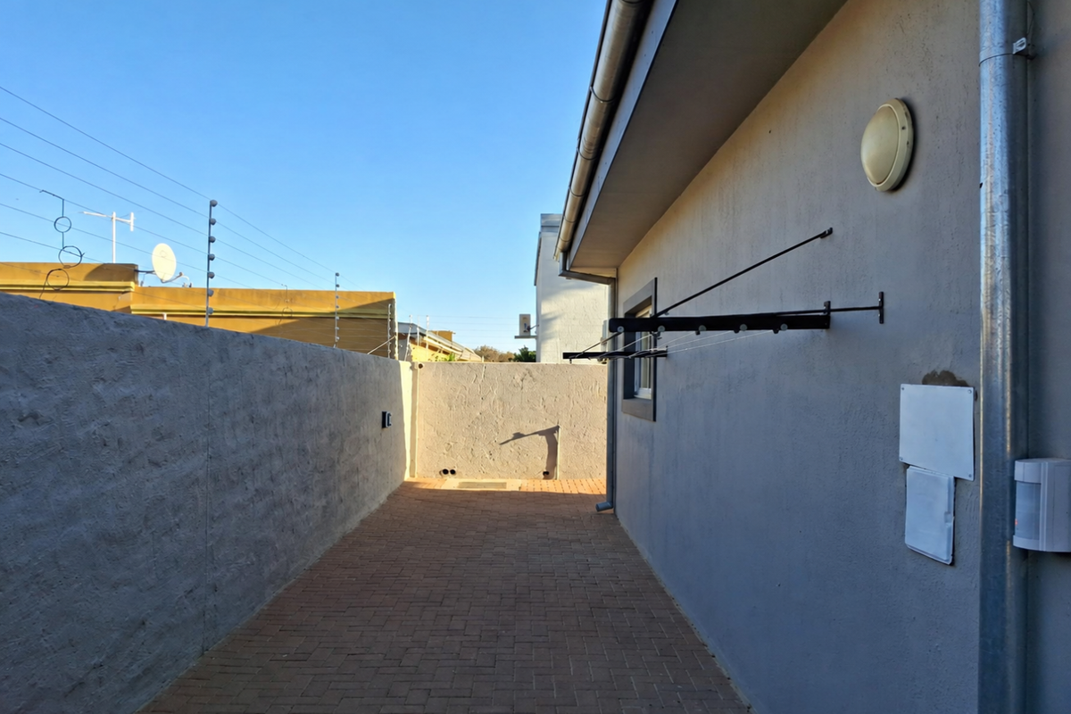 A paved side passage or rear exterior area featuring a boundary wall, wall-mounted clothesline, and exterior security lighting.