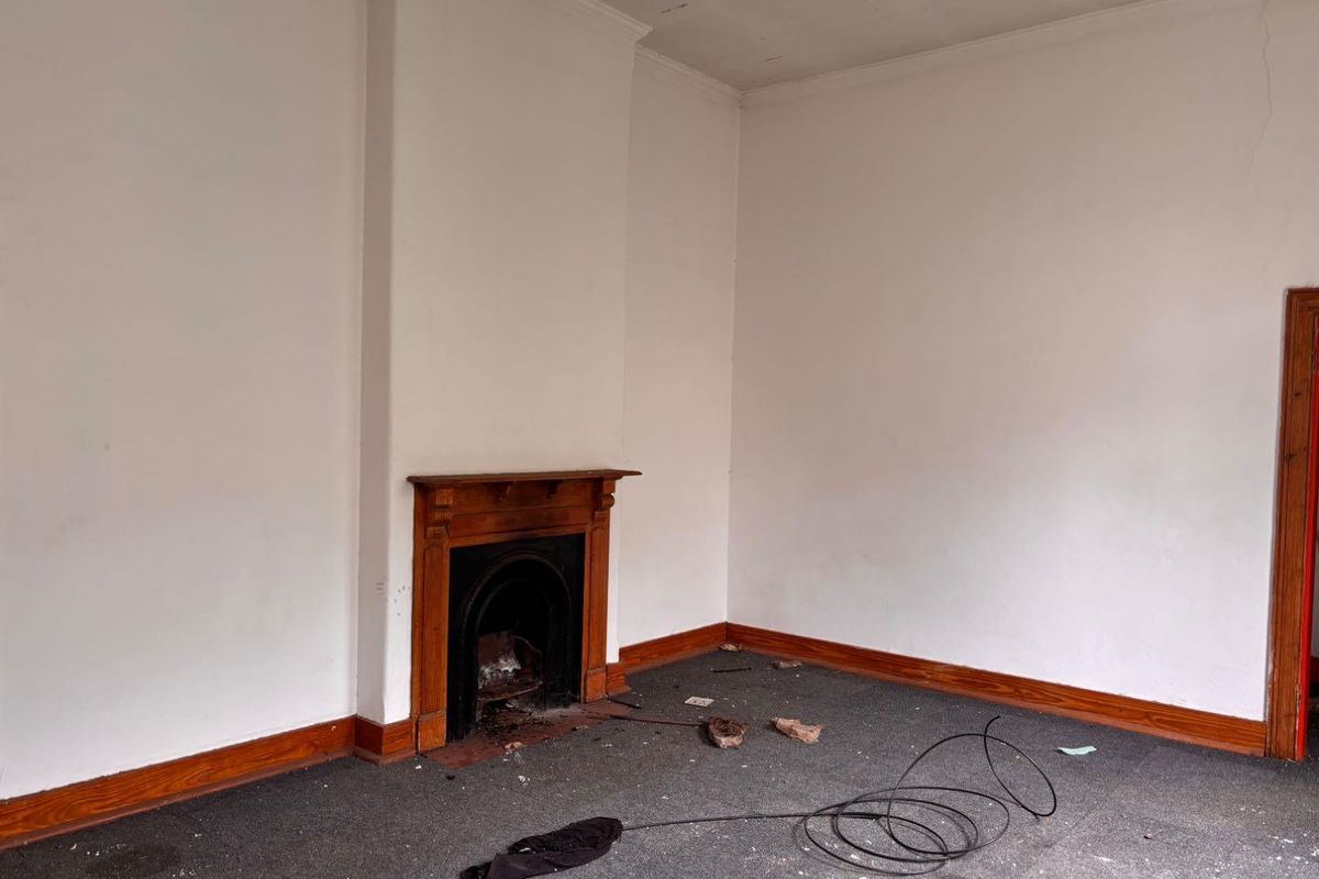 An empty living room featuring a traditional wooden fireplace mantel, timber skirting boards, and carpeted flooring, requiring cosmetic updates.