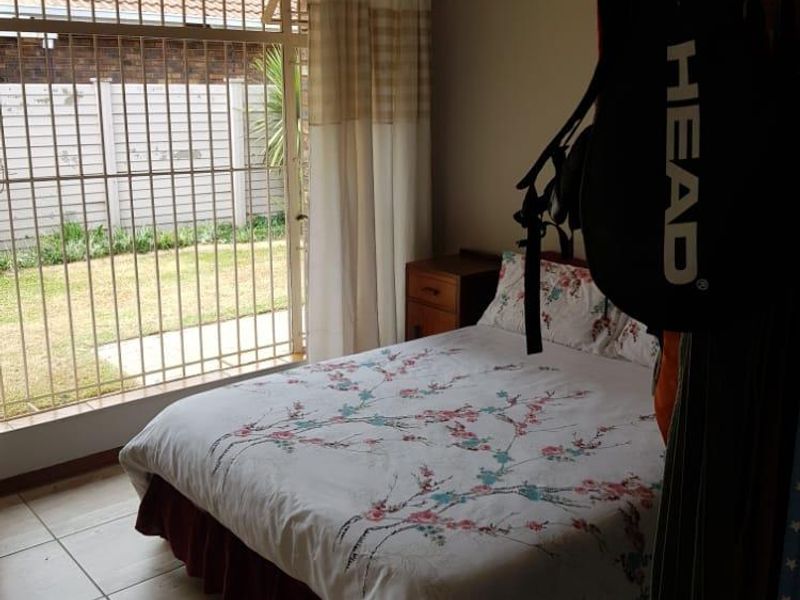 A bedroom featuring tiled flooring, a ceiling fan, and large windows with security bars providing access to the garden area.