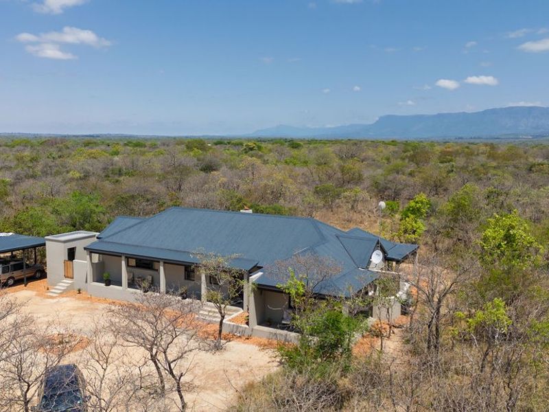 Aerial view of a modern house with a metal roof, featuring a covered carport and expansive natural bushveld surroundings with mountain views.