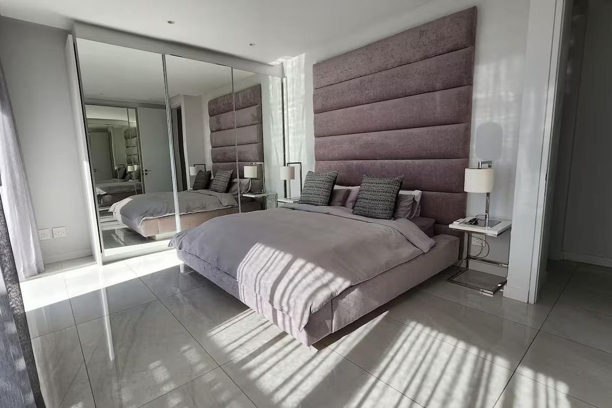Modern bedroom featuring large format floor tiles, a floor-to-ceiling mirrored built-in wardrobe, and a custom upholstered feature headboard.