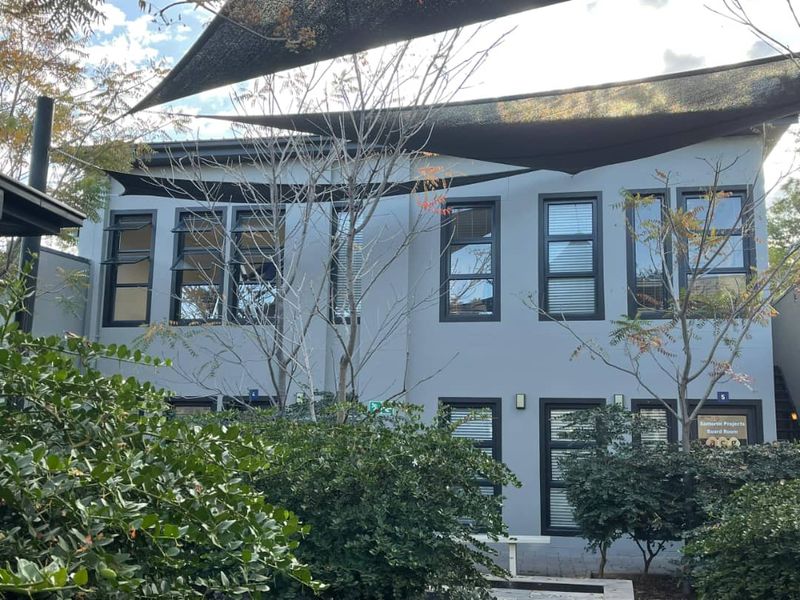 Rear exterior view of the office building featuring modern grey facade, dark aluminium window frames, and overhead shade sails providing outdoor coverage.