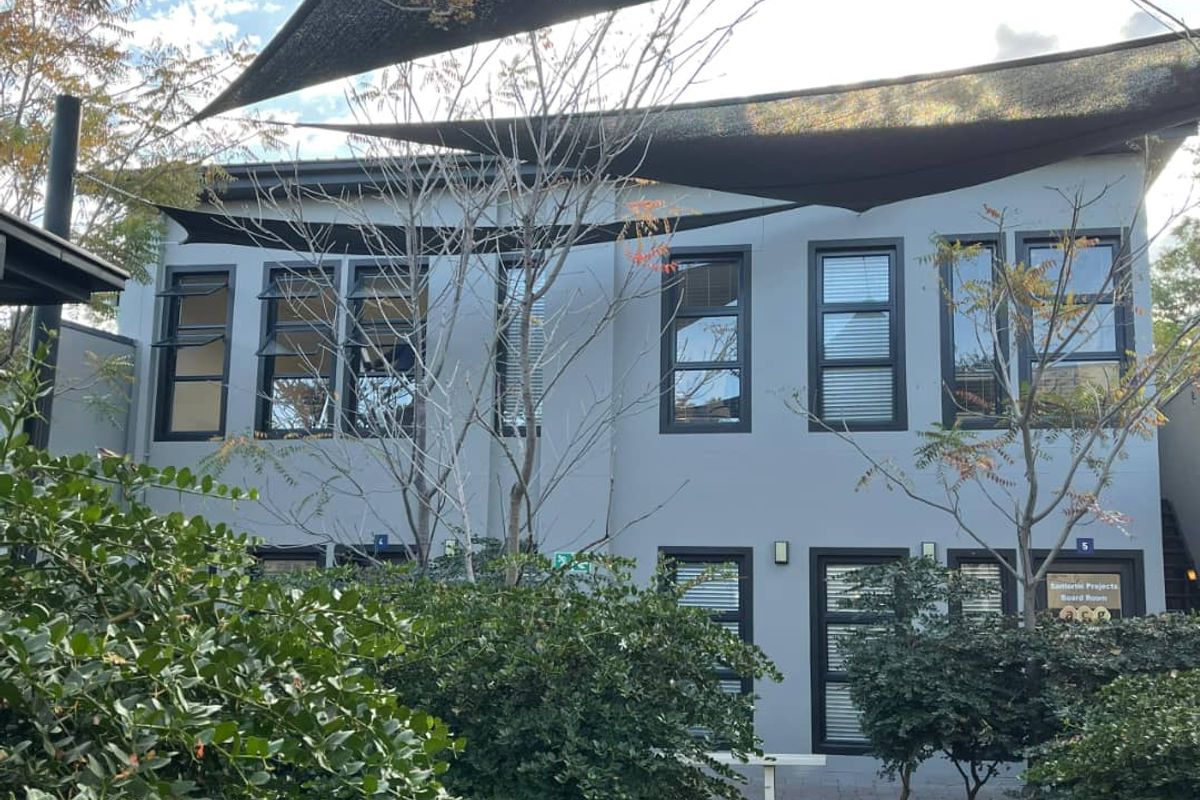 Rear exterior view of the office building featuring modern grey facade, dark aluminium window frames, and overhead shade sails providing outdoor coverage.