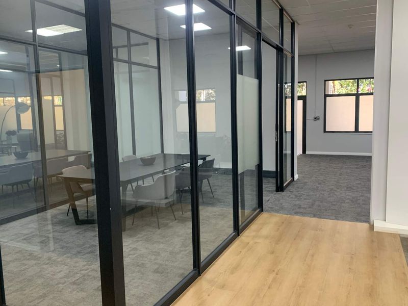 Modern office space featuring glass-partitioned meeting rooms with black aluminum frames, recessed lighting, and a combination of carpet and wood-look flooring.