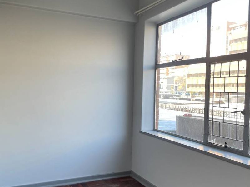 An empty bedroom featuring original parquet flooring, a large steel-framed window with security bars, and neutral wall finishes.