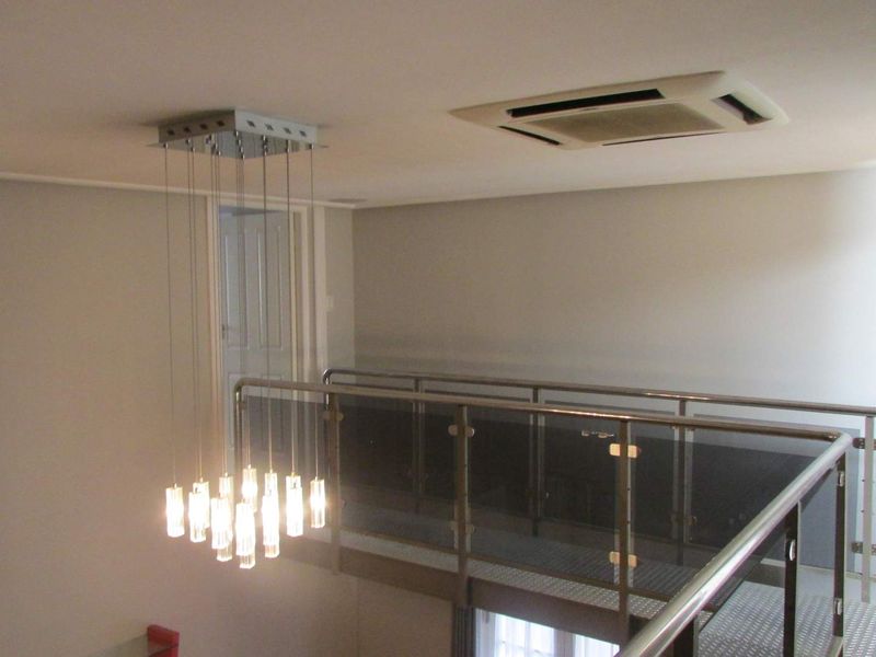 A mezzanine hallway featuring a modern glass and metal balustrade, recessed ceiling air conditioning, and a contemporary multi-pendant light fixture.