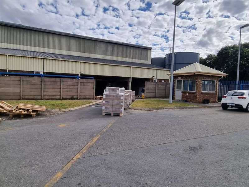Commercial property exterior featuring a large warehouse structure, paved yard area, security guard house, and perimeter fencing.